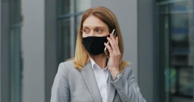 Maskeli beyaz bir kadın sokakta yürüyor ve cep telefonuyla konuşuyor. Solunum korumalı bir kadın yaya dışarıda geziniyor ve cep telefonuyla konuşuyor. Konuşma.
