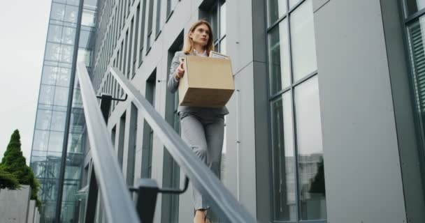 Kaukázusi feldúlt üzletasszony sétál le a lépcsőn egy doboz cuccal, ahogy elhagyja az üzleti központot. A női irodai munkás elvesztette az állását. A munkanélküliségi ráta növekszik a vírus kitörésekor. Kirúgott nő — Stock videók