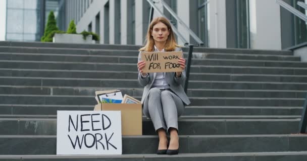 白人忧心忡忡的年轻妇女绝望地坐在城市的楼梯上，拿着海报需要工作和意志为食物而工作。无工作的女性看上去忧心忡忡.封锁后的失业情况 — 图库视频影像