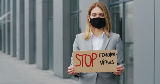 Portrait shot of Caucasian young businesswoman in mask standing outdoor at big stairs and showing table with words Stop Coronavirus. Female demonstrating board with protest against pandemic. — Stock Video