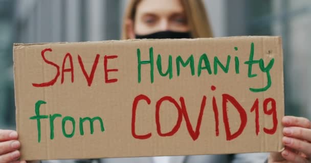 Primer plano de la mesa de cartón con palabras Save Humanity de Covid 19 en manos de una joven caucásica enmascarada. Mujer borrosa mostrando tablero. Protesta solitaria al aire libre. — Vídeos de Stock