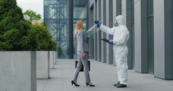 Trabajadora de oficina en máscara yendo a trabajar y hombre en traje blanco y respirador midiendo su temperatura, dejándola ir. Permitir pasar al centro de negocios para el visitante. pandemia de Covid-19. Permiso para entrar. — Vídeos de Stock