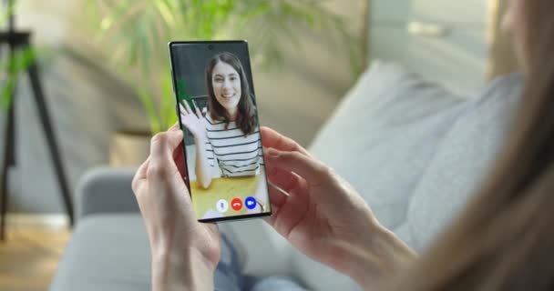 Pantalla del teléfono celular con videollamada entre dos chicas. Mujer teniendo chat de vídeo en línea con bastante chica caucásica en el interior. Mujer hablando a través de la cámara del teléfono inteligente mientras está sentado en la habitación. Concepto de conversación — Vídeos de Stock