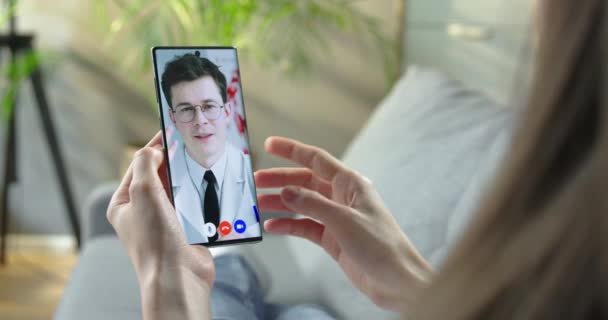Wanita yang berkonsultasi secara online dengan dokter Kaukasia melalui konferensi video di smartphone di rumah. Gadis berbicara dengan dokter di ponsel saat beristirahat di kamar. Konsep layanan kesehatan. — Stok Video