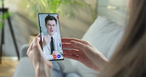 Mujer hablando con el médico en el chat de vídeo en el teléfono celular mientras descansa en la habitación. Mujer que tiene consulta médica en línea con el médico caucásico en videoconferencia sobre smartphone en el hogar Concepto de salud — Vídeos de Stock