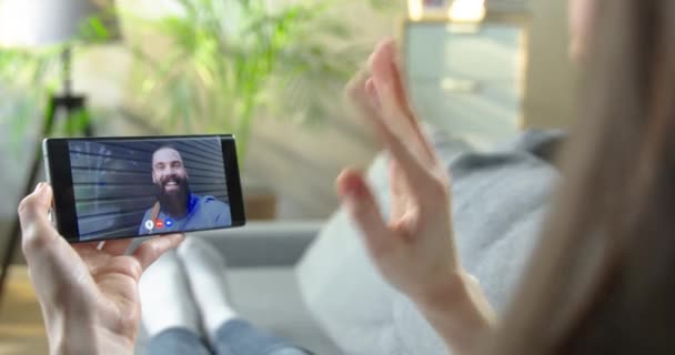 Dívka má videokonferenci na smartphonu s bělošským přítelem, zatímco odpočívá v obývacím pokoji. Žena mluví v online chatu se šťastným vousatým mužem, který mává rukou. Konverzace koncepce — Stock video