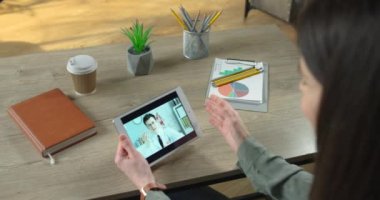 Odada oturan beyaz bir erkek doktorla tablette video sohbeti yapan bir kızın omuz manzarası. Doktor, içeride masa başında oturan bir kadına video konferansı yoluyla sağlık tavsiyesi veriyor..