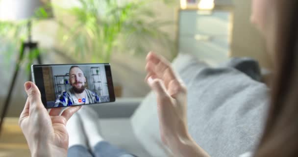Девушка с видеозвонком на смартфон с красивым белым другом во время отдыха в гостиной. Женщина разговаривает в чате со счастливым бородатым мужчиной и машет рукой. Онлайн чат — стоковое видео