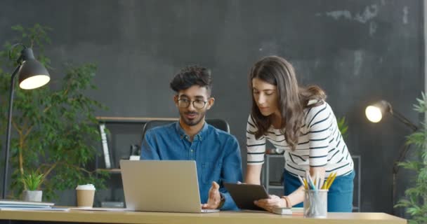 Jovens mestiços homens e mulheres colegas de trabalho conversando alegremente e discutindo o trabalho no escritório. árabe cara sentado na mesa com laptop computador e caucasiano menina de pé ao lado dele com tablet dispositivo. — Vídeo de Stock