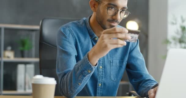 テーブルに座って、クレジットカードを保持し、ラップトップコンピュータ上でオンラインショッピング眼鏡でアラブの若い男性のオフィスの労働者。インターネットで購入し、ロフトキャビネットで支払うハンサムなアラビア人の男. — ストック動画