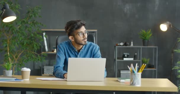 Jovem árabe ocupado trabalhador do escritório masculino sentado à mesa, trabalhando no computador portátil e considerando. Empresário árabe atraente digitando no teclado e mensagens de texto enquanto pensa no gabinete. Café na mesa — Vídeo de Stock