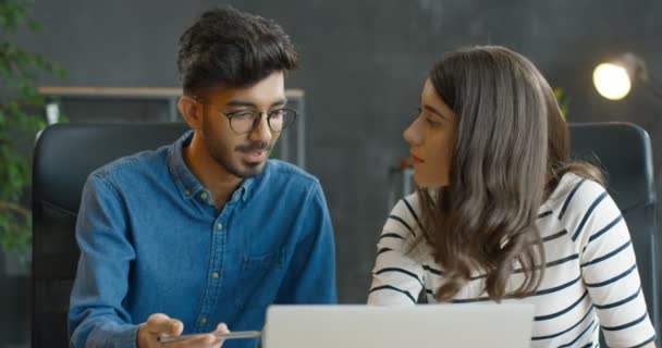 Muliethnic joven chico y chica sentados en la mesa juntos en el ordenador portátil en la oficina y trabajando en el nuevo proyecto de inicio. Trabajadores de empresas mixtas, hombres y mujeres, lluvia de ideas y charlas. — Vídeos de Stock