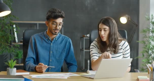 Молодой араб в очках и белая девушка, сидящая за столом в офисе и работающая вместе над стартап-проектом. Коллеги по смешанным расам мужчины и женщины обсуждают работу и проверяют документы. — стоковое видео