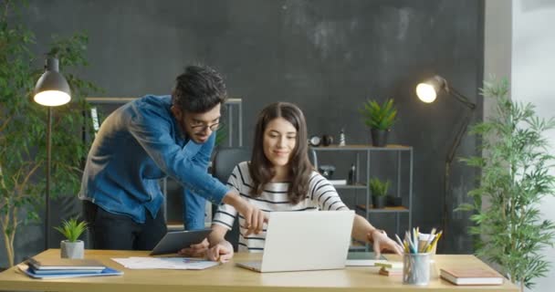 Giovani colleghi multietnici di sesso maschile e femminile che parlano, fanno brainstorming e discutono di lavoro in ufficio. Ragazza caucasica seduta alla scrivania con computer portatile e ragazzo arabo in piedi con dispositivo tablet come aiuto. — Video Stock