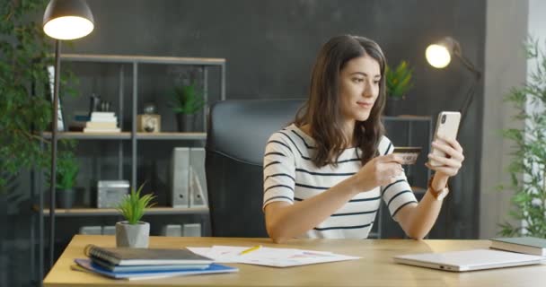Caucasian young female office worker sitting at desk, holding credit card and shopping online on mobile phone. Beautiful girl buying in internet and paying in loft cabinet. — Stock Video