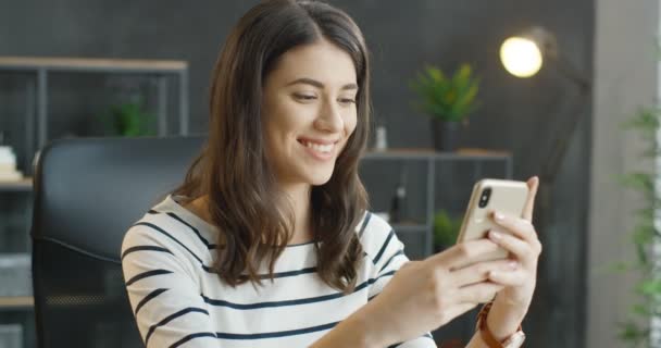 Caucasienne belle femme brune dans des lunettes tenant smartphone dans les mains. Jolie jeune fille souriante tapant et défilant sur le téléphone mobile dans le bureau. Message texte féminin et saisie sur téléphone portable. — Video