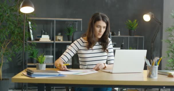 Genç beyaz iş kadını ofiste oturuyor ve dizüstü bilgisayarla ilgili notlar yazıyor. Esmer kadın kontratı kontrol edip okuyor ve planlama yapıyor. İş planlaması — Stok video