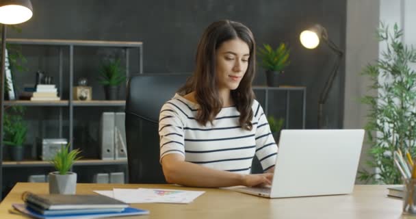 Молода кавказька зайнята жінка, яка сидить за столом, працює над ноутбуком і обдумує питання. Приваблива жінка з брюнеткою друкує на клавіатурі і смс, думаючи в кабінеті. — стокове відео