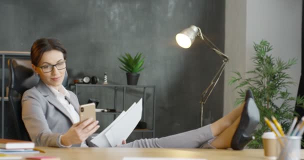 Kaukasiska vackra affärskvinna sitter i skåp, hålla ben på bordet och läsa kontrakt. Kvinnliga kontorsarbetare kontrollera dokument och smartphone meddelande. Kvinnan avlyssnar telefonen och vilar. — Stockvideo