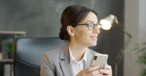 Kavkazská krásná brunetka obchodnice v brýlích drží smartphone v rukou. Pěkně usměvavá žena poklepávala na mobilní telefon v kanceláři. Ženské textové zprávy a psaní na mobilu. — Stock video