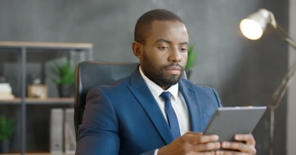 Africano americano empresário sério segurando e usando dispositivo tablet. Bonito homem batendo e rolando no computador no escritório. Mensagens masculinas e digitação na tela e pensamento. Trabalhando no gabinete. — Vídeo de Stock