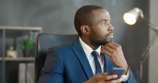 Afrikansk amerikansk seriös affärsman som håller och använder smartphone. Snygg man knackar och bläddrar på mobiltelefon på kontoret. Man sms: ar och tänker. Arbete i kabinett. — Stockvideo