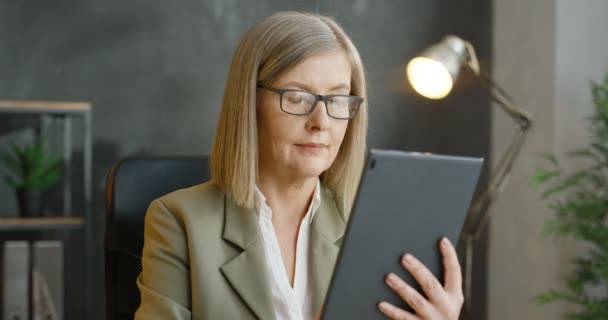 Kaukasiske forretningskvinde holder og bruger tablet enhed i hænderne. Smuk senior kvinde i briller trykke og rulle på computeren på kontoret. Kvindelig sms 'e og skrive og tænke i skab. – Stock-video