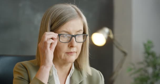 Portrait d'une femme d'affaires blonde caucasienne senior enlevant des lunettes, regardant du côté et pensant. À l'intérieur. Gros plan sur le visage de la femme sérieuse fatiguée d'âge moyen au bureau. — Video