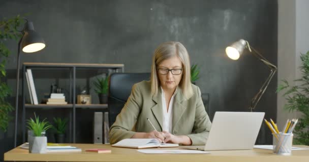 白种人高级女商人坐在办公室，用笔记本写便条，在笔记本电脑上工作。中年妇女注意到在规划师。从事商业规划的女上司. — 图库视频影像