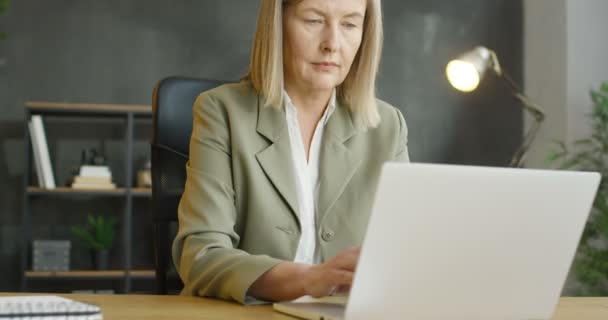 Primer plano de caucásica de mediana edad ocupada trabajadora de oficina sentada en la mesa y trabajando en el ordenador portátil. Hermosa mujer de negocios senior escribiendo en el teclado. Mujer en el gabinete. — Vídeo de stock