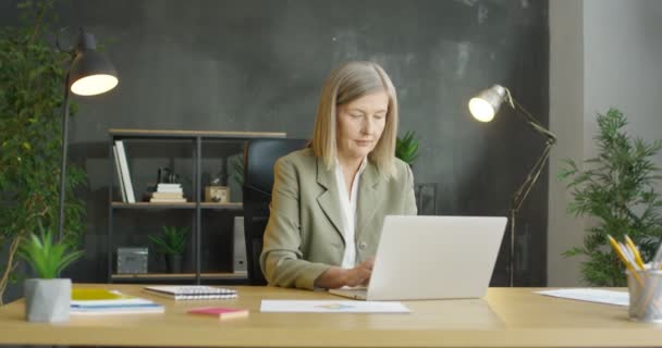 Běloška středního věku, zaměstnaná kancelářská pracovnice, sedí u stolu a pracuje na laptopu. Krásná starší podnikatelka, co píše na klávesnici. Žena ve skříni. — Stock video