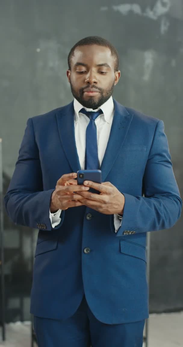 Africano americano feliz homem de negócios em pé no escritório e mensagem de texto no smartphone. Masculino tocando e rolando no telefone celular. Homem de terno e gravata sorrindo alegremente. Tiro vertical para histórias. — Vídeo de Stock