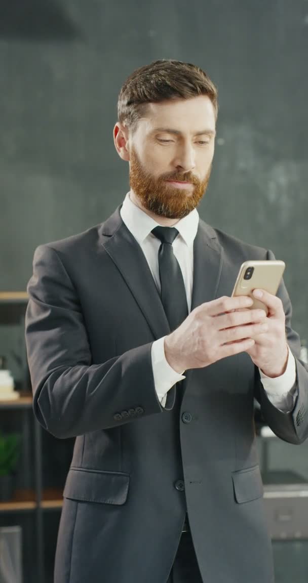 Καυκάσιος νεαρός χαρούμενος επιχειρηματίας στο γραφείο στέλνει μηνύματα στο smartphone. Όμορφος άντρας που πατάει κύλιση στο κινητό τηλέφωνο. Άνδρας με κοστούμι και γραβάτα χαμογελώντας χαρούμενα. Κάθετο υλικό για ιστορίες. — Αρχείο Βίντεο