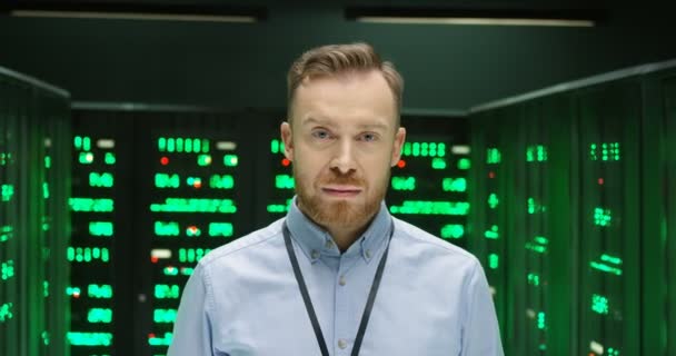 Retrato del joven caucásico guapo mirando directamente a la cámara en la sala de almacenamiento de big data con muchos procesadores y sonriendo. Administrador del sistema masculino de pie entre los servidores en el centro de datos. — Vídeos de Stock