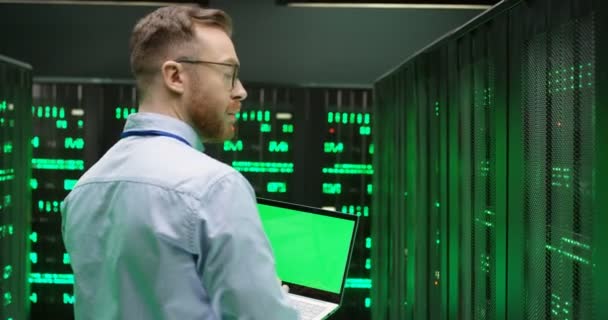 Achter van Kaukasische jongeman in glazen lopen tussen servers met laptop computer van groen scherm en het controleren van het werk van big data processors. Chroma sleutel. Achteraanzicht. Mannelijke analyticus in gegevensopslag. — Stockvideo