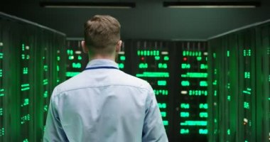 Beyaz tenli yakışıklı genç adam büyük veri bilgileriyle sunucuların arasında dolaşıyor. Veritabanı deposunda çalışan yeşil işlemciler. Dijital telekomünikasyonda çalışan erkek işçiye arka plan.