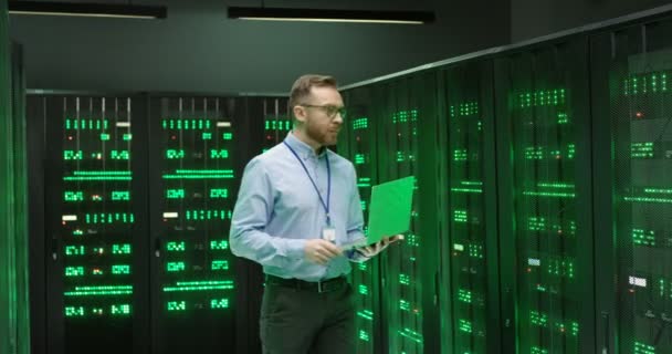 Bělošský mladík v brýlích stojí mezi servery s notebookem, píše na klávesnici a kontroluje velké datové procesory. Mužská analytika v ukládání dat při práci na zabezpečení digitálních informací. — Stock video