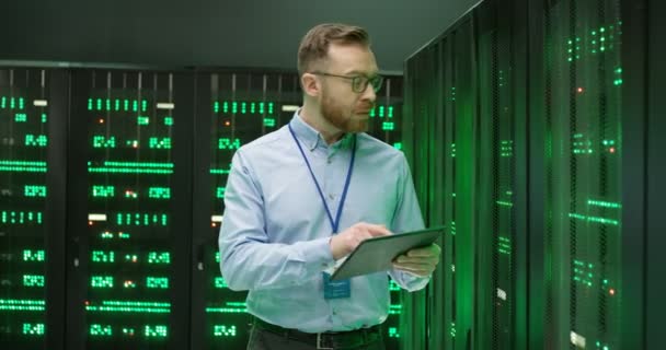 Bělošský mladík v brýlích stojí mezi servery s tabletovým zařízením, ťuká na obrazovku a kontroluje velké datové procesory. Mužská analytika v ukládání dat při práci na zabezpečení digitálních informací. — Stock video