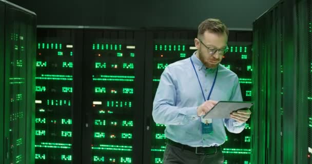 Homem jovem caucasiano em óculos andando em servidores com dispositivo tablet, tocando e verificando processadores de big data. Analítico masculino no armazenamento de dados trabalhando na segurança da informação digital do banco de dados. — Vídeo de Stock