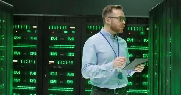 Bělošský mladík v brýlích kráčející na serverech s tabletovým zařízením, ťukající a kontrolující velké datové procesory. Mužský inženýr v ukládání dat pracuje na zabezpečení digitálních informací v databázi. — Stock video