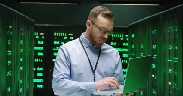 Hezký běloch v brýlích, stojící ve velkém úložišti dat u procesorů, píší na klávesnici laptopu a pracuje na zabezpečení databáze. Mužská analytika v datacentru. — Stock video