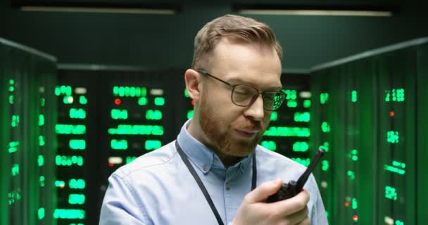 Detailní záběr mladého bělocha v brýlích, jak mluví vysílačkou v databázovém úložišti. Muž bezpečnostní pracovník datacentra a telekomunikačních serverů místnosti. — Stock video