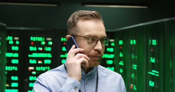 Běloch v brýlích stojí v databázové místnosti se servery, mluví na mobilním telefonu a kontroluje zabezpečení dat. Detailní záběr mužské analytiky a inženýra v datacentru mluvící na mobilním telefonu. — Stock video