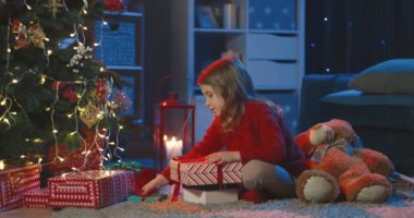 Kafkasyalı küçük bir kız Noel ağacında yerde oturuyor ve bayram akşamı karanlık odada hediyeler katlıyor..