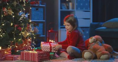 Kırmızı kazaklı, neşeli beyaz kızın portresi Noel ağacındaki hediyelere bakıyor ve gülüyor..