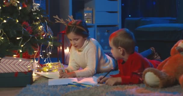 照片中的白种人可爱的小男孩和小女孩躺在圣诞树下的地板上，一边聊天，一边画画。晚上在家里. — 图库视频影像