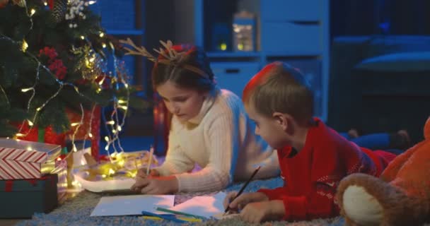 Piccoli bambini carini caucasici sdraiati sul pavimento sotto l'albero x-mas, parlando allegramente e disegnando. Di notte.. — Video Stock