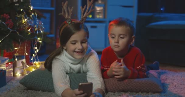 Närbild av den lilla glada syster och bror som ligger på golvet vid julgranen och titta på karikatyrer på smartphone i det mörka rummet. — Stockvideo