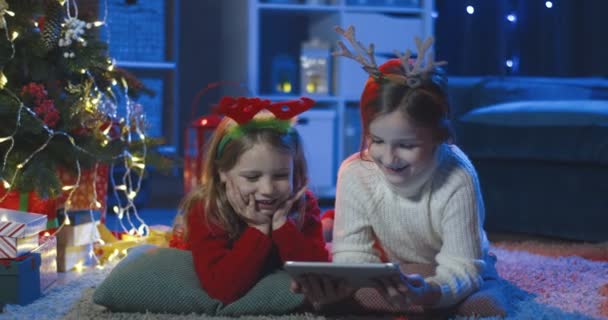 Närbild av de små glada och vackra systrar som ligger på golvet i x-mas dekorerade mörka rum och titta på karikatyrerna på surfplattan datorn. Porträtt. — Stockvideo