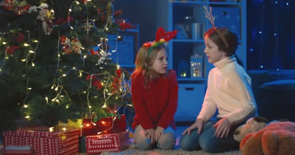 Zwei kleine kaukasische Mädchen mit den lustigen Ohren auf dem Kopf sitzen abends am Weihnachtsbaum und unterhalten sich. Ferienzeit. — Stockvideo